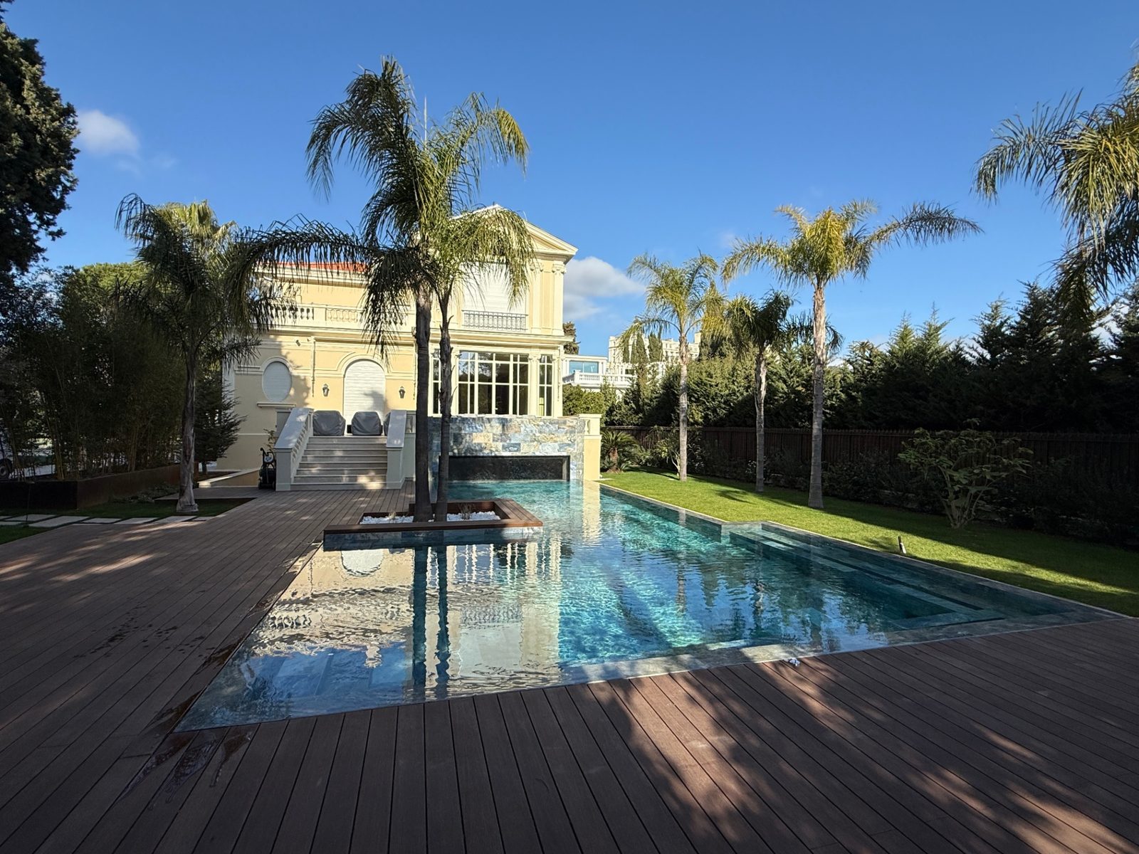 Piscine miroir haut de gamme à débordement périphérique, plage en bois et palmiers devant une villa